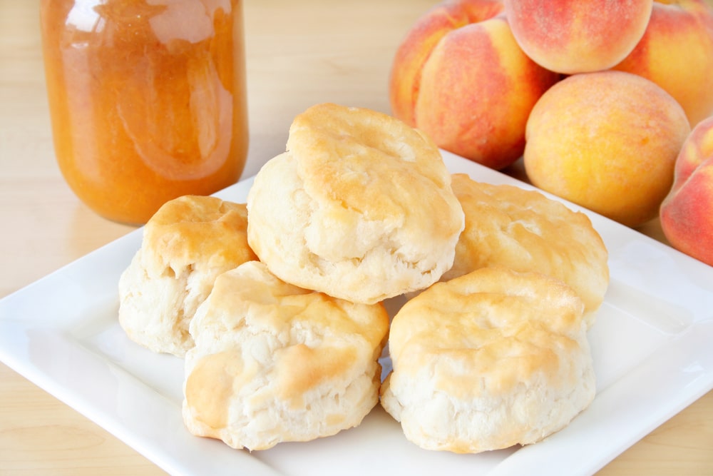 Freshly baked biscuits stacked on a white plate, accompanied by homemade peach preserves and a selection of fresh peaches.