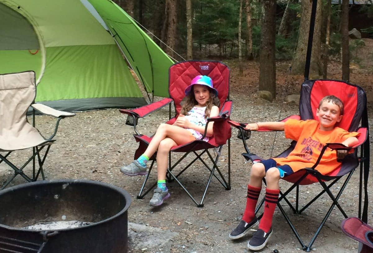 Two cheerful children seated on camping chairs in front of a green tent in a forested area