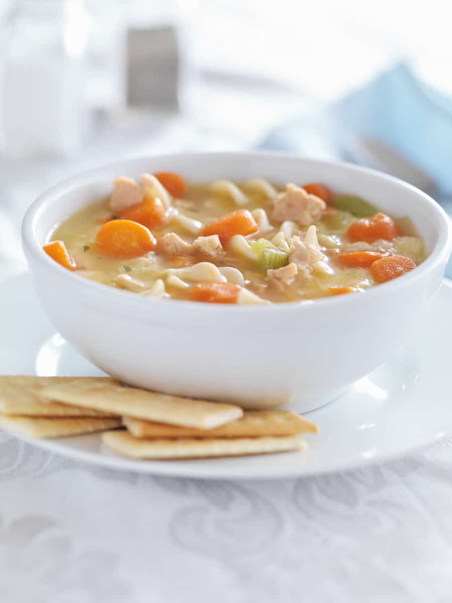 White bowl of hearty chicken noodle soup, accompanied by a side of crackers.