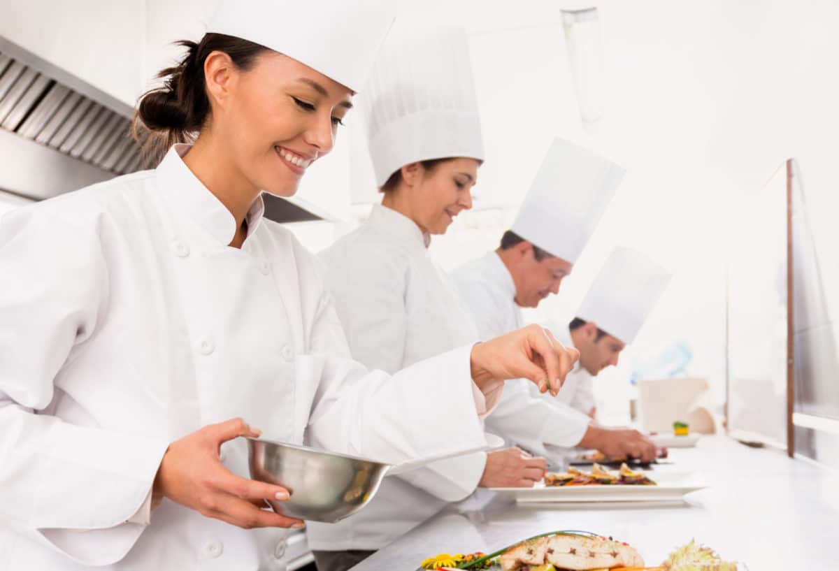 Line of professional chefs in white attire actively cooking and preparing dishes in a modern kitchen.