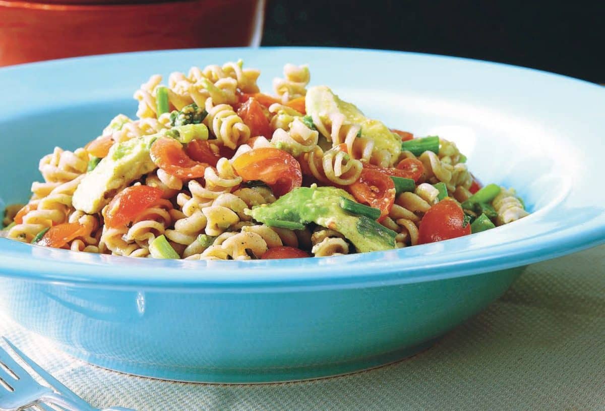 Blue bowl filled with pasta salad containing fresh tomatoes, avocado, and other vegetables.