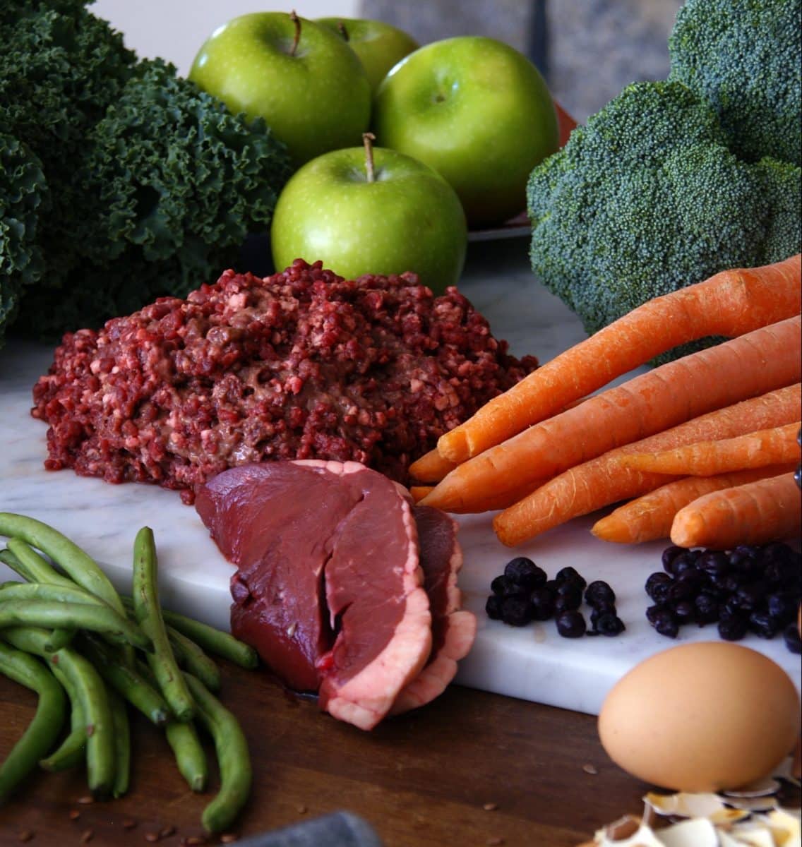 Assortment of raw ingredients including ground meat, green apples, broccoli, carrots, egg, and green beans on a counter.