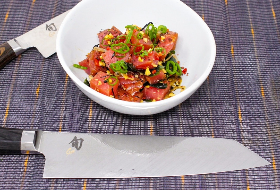 A white bowl containing Hawaiian-style Ahi Poke, flanked by two Shun knives on a blue tabletop.