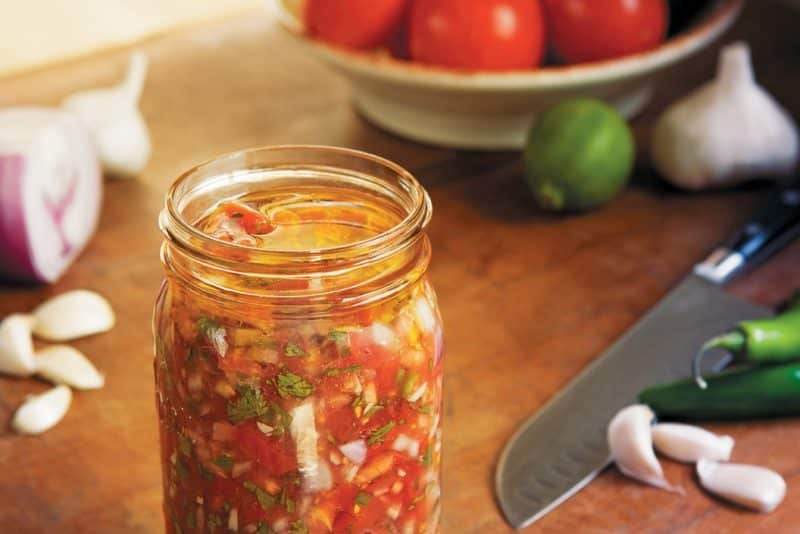 A jar filled with vibrant and spicy fermented summer salsa, surrounded by fresh ingredients.