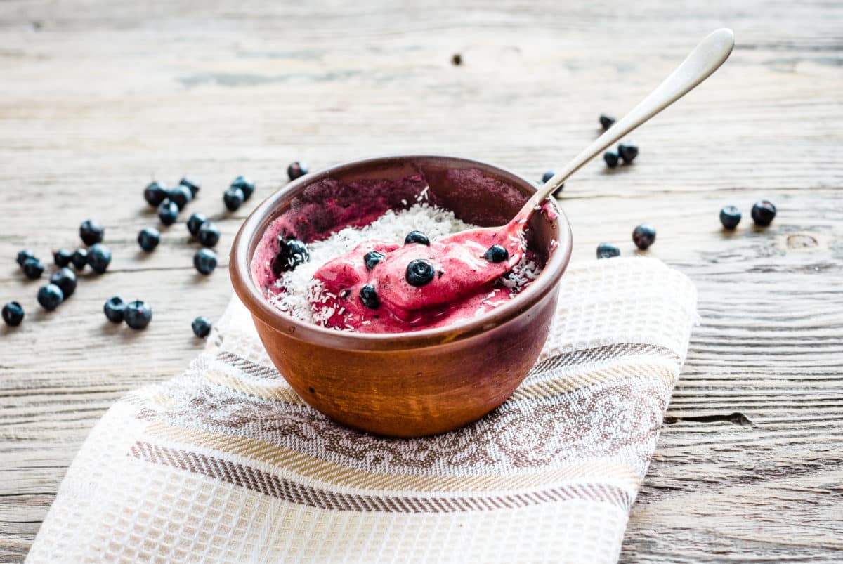 A bowl of vegan banana ice cream topped with fresh blueberries and coconut shavings, placed on a rustic wooden table.